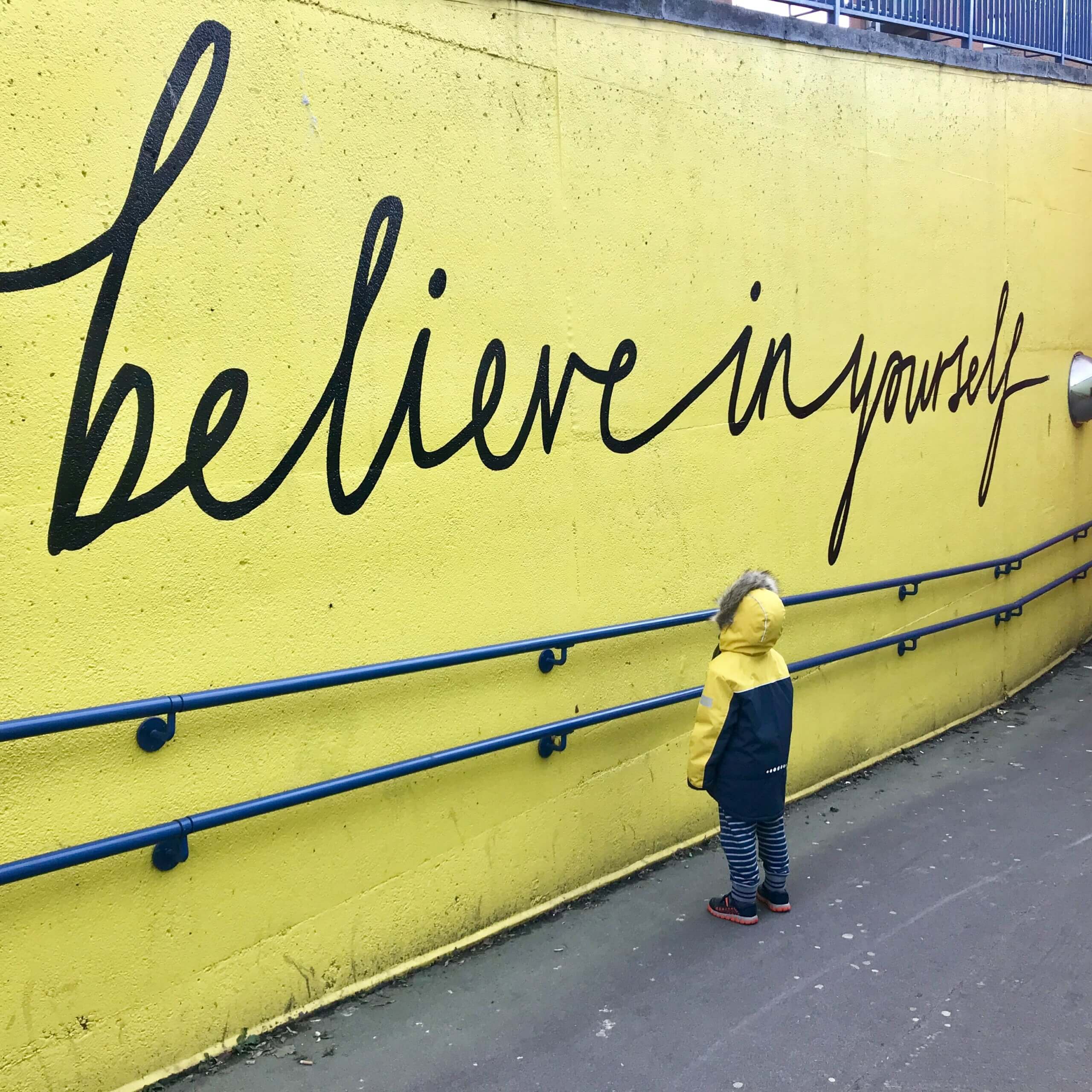 Ein Kind in gelber Jacke steht vor einer gelben Wand, auf der "Believe in yourself" steht