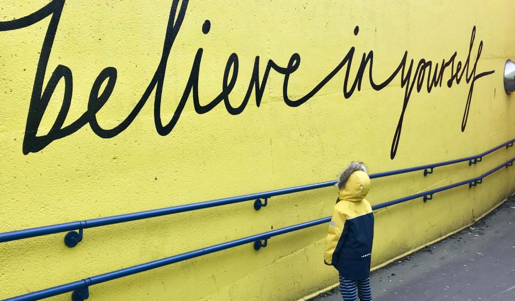 Ein Kind in gelber Jacke steht vor einer gelben Wand, auf der "Believe in yourself" steht